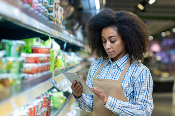 A grocery store employee in an apron checks the inventory on the shelves using a tablet. The worker...