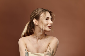 A young woman with vitiligo and a tattoo on her arm poses with her shoulders bare against a beige background.