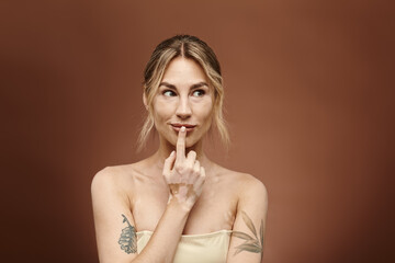 A young woman with vitiligo poses with her shoulders bare, her finger to her lips, looking off to the side.