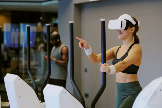 Young woman playing VR game and smiling widely while she doing cardio, African American man surfing Internet in background