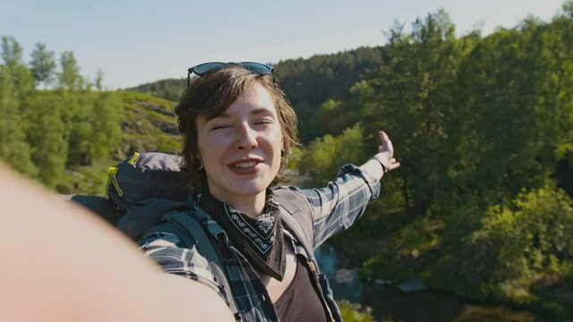 Handheld POV shot of young happy Caucasian female tourist speaking at camera while vlogging from outdoor summer hiking journey, standing against beautiful landscape with stream and forest