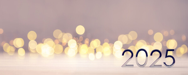 Metal numbers 2025 on a white table with Christmas trees and bokeh lights. Happy New Year 2025 is...