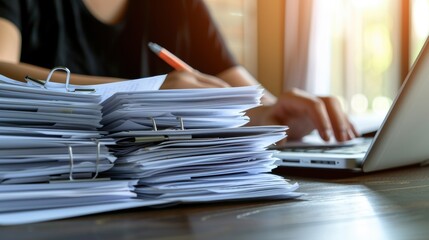 A large stack of documents on a desk with a person working on a laptop in the background.