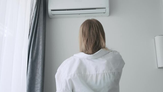 Back view of exhausted sweaty woman turning on an air conditioner with a remote control to cooling off during hot summer weather, suffering from heat, high temperature
