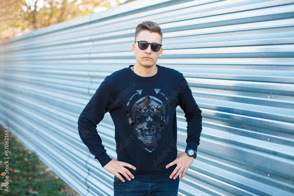 Poster Handsome young man model with hairstyle with fashion sunglasses in black pullover near metal background on the street