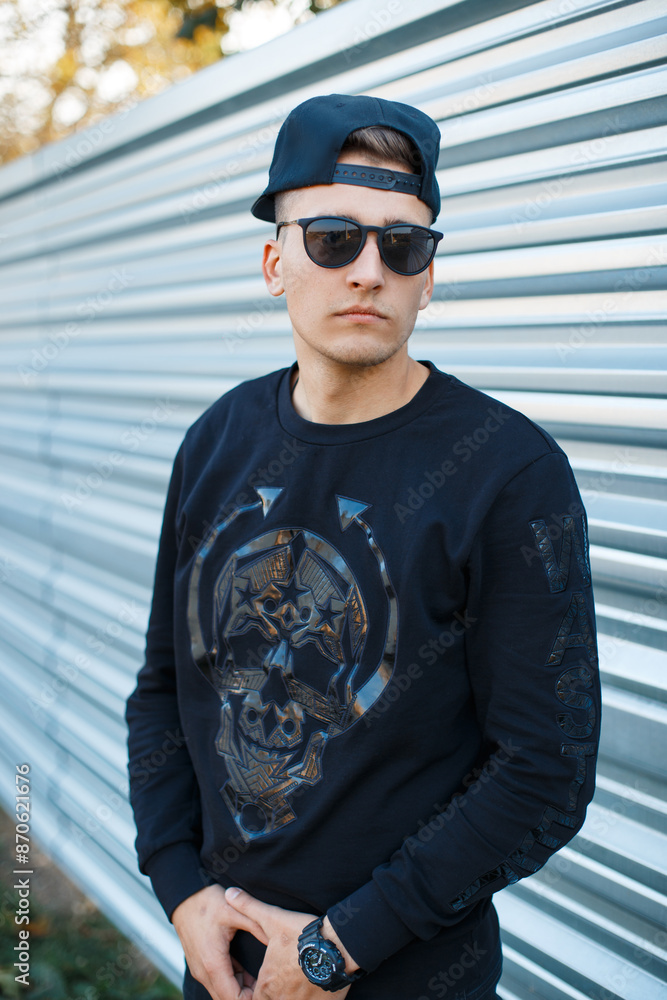 Wall mural Fashion handsome young hipster man with stylish sunglasses with a baseball cap in a fashionable black pullover on the street near a metal fence