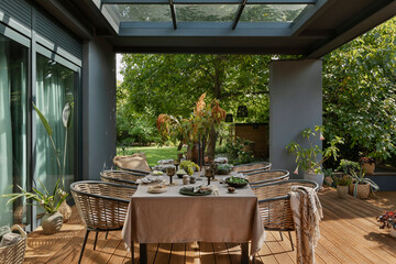 Creative composition of warm and cozy terrace with table, garden furnitures, vase with flowers, wooden floor, plaid, curtain, grape, big garden and personal accessories. Home decor. Template.