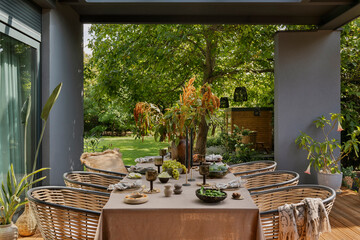 Creative composition of warm and cozy terrace with table, garden furnitures, vase with flowers, wooden floor, plaid, curtain, grape, big garden and personal accessories. Home decor. Template.