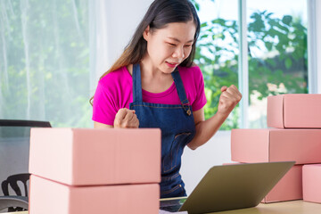 A woman in overalls sells products online, inspects parcels, takes customer orders, works at a desk, and uses pink boxes to pack items ready for shipping to customers.