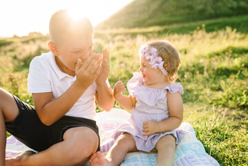 Older brother plays with younger sister in spring sunlight. Kids playing in mountains on summer day. Happy children hug and sit on blanket in green grass in field at sunset. Spending time together.