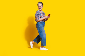 Fototapeta premium Full body photo of attractive retired woman walk hold telephone dressed stylish colorful clothes isolated on yellow color background