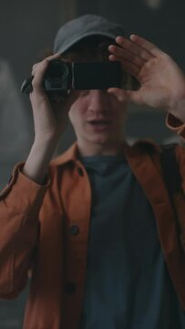 Vertical medium shot of Caucasian guy recording content on video camera, filming interior of abandoned mansion while examining dark room with friends