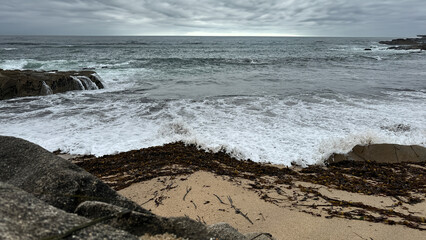 Stormy Ocean Waves Crashing on Rocky Shore with Seaweed and Cloudy Sky.