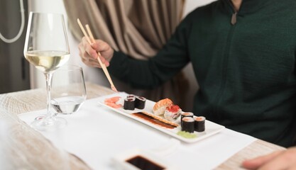 A person enjoys a meal of sushi and white wine at a white-clothed table in a modern setting