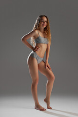 Fototapeta premium Full-length image of beautiful young girl showing her perfect fit body, model posing in underwear against grey studio background. Concept of female beauty, body and skin care, fitness, wellness