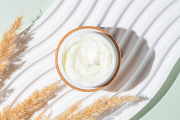 Cosmetic cream with dry pampas flowers on light concrete podium on green background. Dermatologic, components for cosmetics.