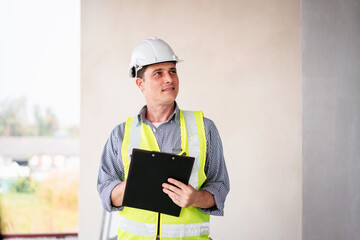 A man wearing a yellow vest and a white helmet is holding a clipboard