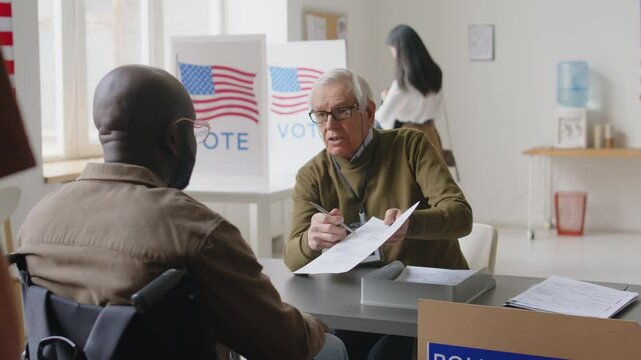 Black man in wheelchair riding to senior Caucasian inspector giving him ballot and explaining how to vote at polling place on Election day