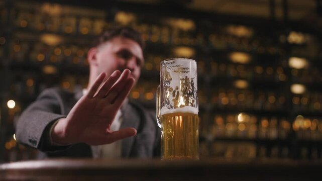 A man refuses to drink beer by extending his hand and showing a stop sign. The focus is on the half-filled beer glass. The man is out of focus