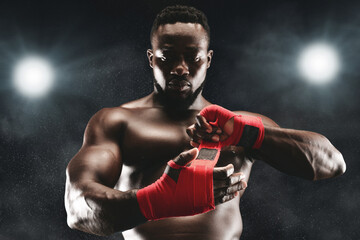 A muscular boxer with a serious expression wraps his hands with red tape in a dimly lit boxing ring.