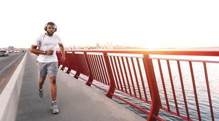 A man in headphones runs on a city bridge with a red railing. The sunset is in the background, casting a warm glow on the scene.