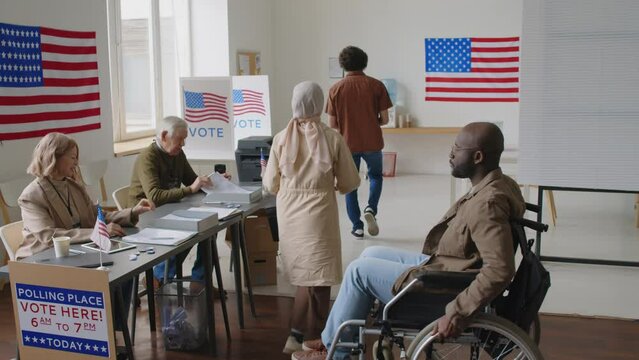 African American man with disability riding wheelchair in polling place, taking ballot from female election inspector and going to vote