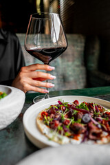 Close-up of a hand holding a glass of red wine, with a focus on the wine’s rich color and the elegant stemware, against a blurred background of gourmet food