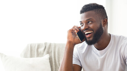 African American young man, wearing a white t-shirt and sitting on a couch at home, is talking on the phone while smiling broadly. The man has a short haircut and a full beard, copy space