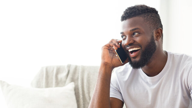 African American young man, wearing a white t-shirt and sitting on a couch at home, is talking on the phone while smiling broadly. The man has a short haircut and a full beard, copy space