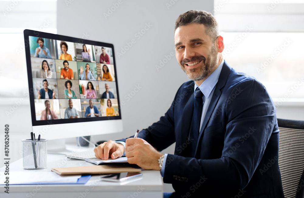 Poster A businessman in a suit sits at a desk in an office, looking at the camera while participating in a video conference meeting on a computer monitor. He is smiling and holding a pen in his hand