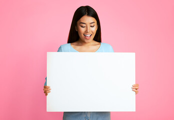 Asian young woman with long, dark hair smiles while holding a blank white sign against a pink background. She is wearing a light blue t-shirt and denim shorts. She is looking down at the sign