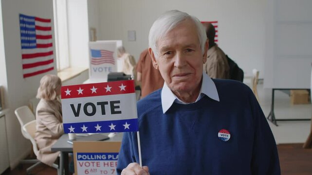 Portrait of elderly man with voter badge and flag posing for camera at polling place on Election day in USA