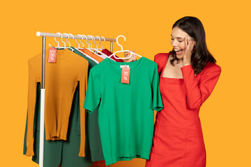 A woman is excitedly looking at a green t-shirt hanging on a clothing rack. She is wearing a red dress and has her hand on her face. There are other clothes hanging on the rack