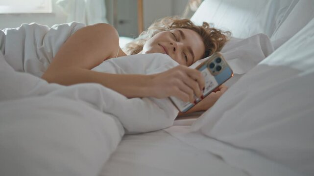 Closeup peaceful woman sleeping in bed. Sleepy curly girl checking smartphone