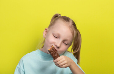 A beautiful joyful girl with white hair of seven years old eats a chocolate bar on a yellow...