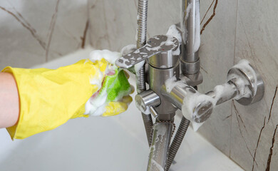 A girl's hand in a yellow rubber glove washes a water tap in the bathroom with a cleaning agent,...