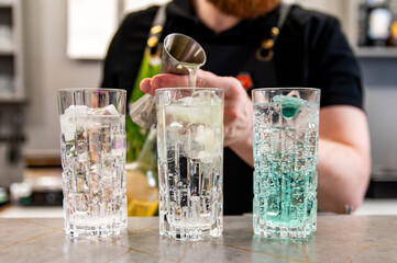 A person in an apron pours liquid into a glass with ice, alongside two other glasses.