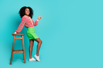 Full length photo of cheerful pretty lady dressed print blouse sitting stool pointing empty space...