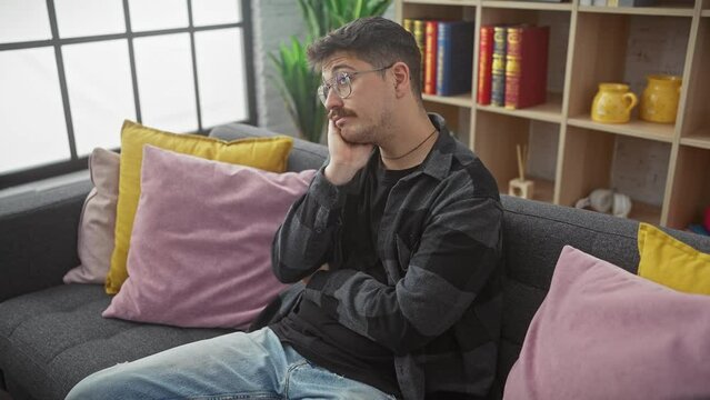 Young hispanic man with glasses, tired and bored, contemplating life&rsquo;s problems on a sofa, crossed arms reflecting depression at home