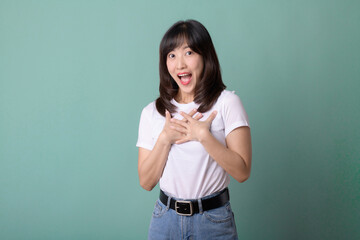 Laughing asian young woman with hands on chest wearing white t-shirt and jeans standing isolated on green background.