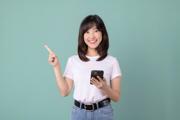 Smiling asian woman pointing while holding smartphone. Woman in white t-shirt and jeans, isolated on green background.