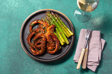 Grilled octopus with asparagus served on a black plate, highlighting vibrant colors and textures against a turquoise background.