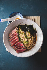 A bowl of ramen featuring perfectly seared tuna slices, garnished with fresh greens, seaweed, and served in a savory broth.