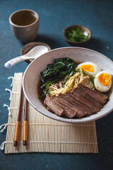 Bowl of ramen with slices of meat, soft-boiled eggs, spinach, and noodles in rich broth, served with chopsticks on a bamboo mat