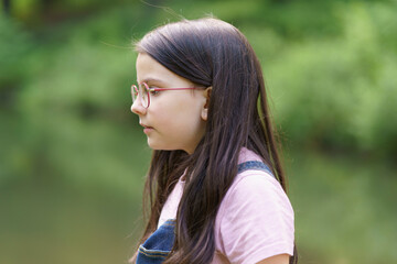 A little cute teenage girl in glasses stands near a pond. Child in denim overalls. Happy childhood...