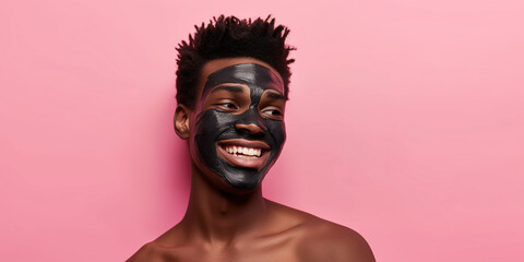 Smiling African-American man with clay facial mask and towel on head uses beauty products and cosmetic on pink studio background. Copy paste empty place for text. Skin care and peeling concept