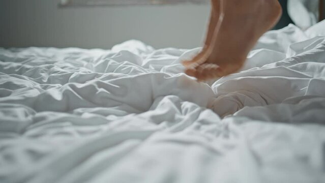Woman legs jumping bed in sunlight closeup. Cheerful lady falling soft pillows