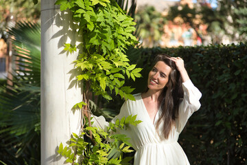 Young woman, natural, with brown hair and white dress, smiling and happy while touching her hair leaning on a marble column. Concept naturalness, simplicity, hairstyles, happiness.