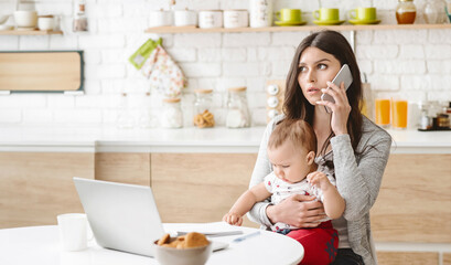A woman sits in her kitchen with a baby in her arms. She is on the phone, looking concerned, with her laptop open in front of her. The kitchen is bright and modern