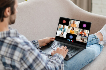 Man sitting on a couch, using a laptop to attend a video conference call. The laptop screen displays several participants in a grid format, indicating that it is an online meeting.